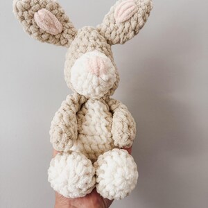May include: A plush crochet rabbit toy crafted from thick, textured beige and cream yarn. The stuffed animal features long ears with soft pink centers, a round cream-colored muzzle with a small pink nose, and a cream belly patch.
