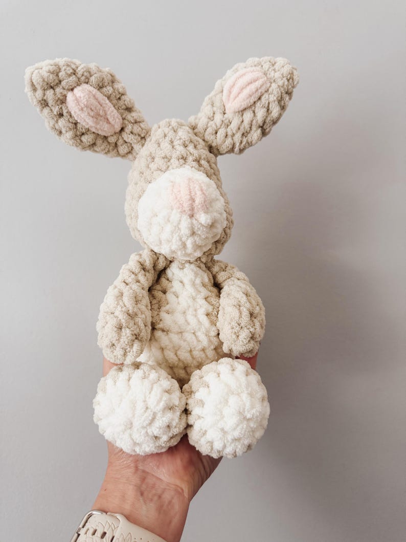 May include: A plush crochet rabbit toy crafted from thick, textured beige and cream yarn. The stuffed animal features long ears with soft pink centers, a round cream-colored muzzle with a small pink nose, and a cream belly patch.