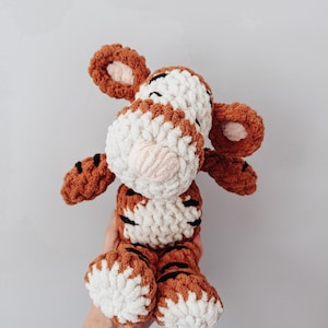 May include: A plush tiger toy made of chunky, soft yarn in shades of burnt orange, cream, and white. The stuffed animal features a striped pattern, rounded ears with cream centers, and a prominent cream-colored muzzle with a small pink nose.
