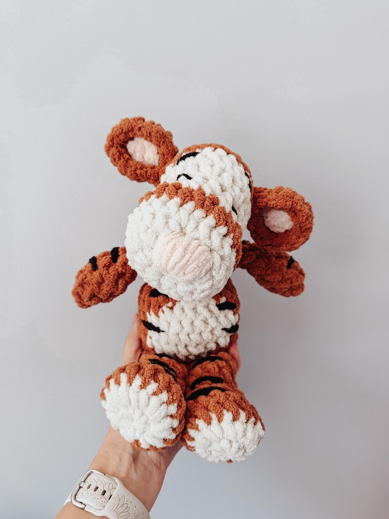 May include: A plush tiger toy made of chunky, soft yarn in shades of burnt orange, cream, and white. The stuffed animal features a striped pattern, rounded ears with cream centers, and a prominent cream-colored muzzle with a small pink nose.
