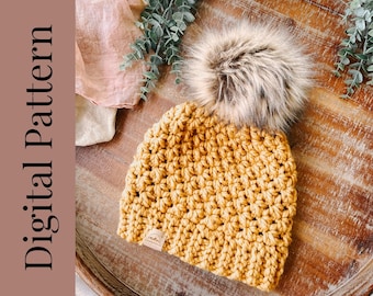 Op de afbeelding: Een gebreide beanie muts in een mosterdgele kleur met een nep bont pompon bovenop. De muts ligt op een houten oppervlak.