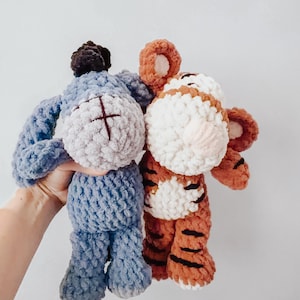 May include: Two handmade crochet plush toys featuring a blue donkey with a dark mane and a striped orange and white tiger. Both stuffed animals are crafted from soft, textured yarn with embroidered facial details and rounded, plush limbs.