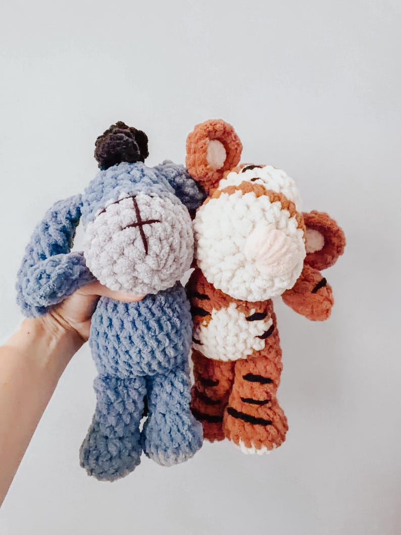 May include: Two handmade crochet plush toys featuring a blue donkey with a dark mane and a striped orange and white tiger. Both stuffed animals are crafted from soft, textured yarn with embroidered facial details and rounded, plush limbs.