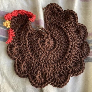 May include: A brown crocheted chicken with a red and yellow comb. The chicken is made with a textured stitch and has a scalloped edge.