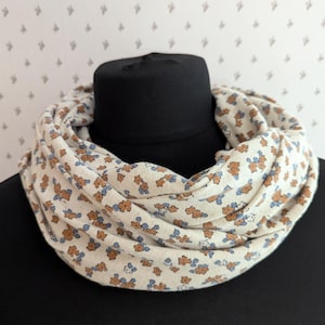 Könnte beinhalten: Ein cremefarbener Loop-Schal mit einem sich wiederholenden Blumenmuster in Braun und Blau. Der Schal ist um einen schwarzen Schaufensterkopf drapiert, was sein kreisförmiges Design und seine weiche Textur hervorhebt. Der Hintergrund ist eine hellfarbige Wand.