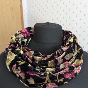 Könnte beinhalten: Ein schwarzer Loop-Schal mit einem floralen Muster in Rosa, Grün und Gold. Der Schal aus weichem, samtartigem Material ist um einen schwarzen Mannequinkopf drapiert. Der Hintergrund ist eine helle Wand mit einem dezenten Muster.