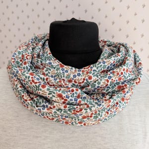 Könnte beinhalten: Ein floraler Rundschal mit einem sich wiederholenden Muster aus roten, blauen und grünen Blumen und Blättern auf weißem Grund. Der Schal ist um einen schwarzen Mannequinkopf gewickelt und zeigt sein kreisförmiges Design.
