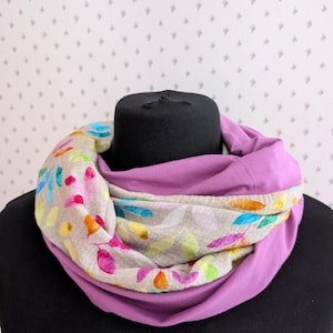 Könnte beinhalten: Ein lila-farbener Loop-Schal mit einem bunten Blumenmuster. Der Schal wird auf einem schwarzen Schaufensterpuppenkopf präsentiert. Das Blumenmuster enthält rosa, orange, gelbe, blaue und grüne Akzente.