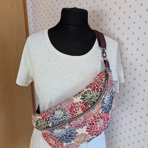 Könnte beinhalten: Eine Bauchtasche mit floralem Muster und geschwungener Form, mit Reißverschluss und burgunderfarbenem Riemen. Das Muster umfasst rote, blaue und grüne Blumen auf cremefarbenem Hintergrund. Die Tasche wird auf einer Schaufensterpuppe präsentiert.