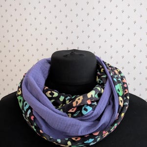Könnte beinhalten: Ein schwarzer und violetter Loop-Schal mit einem farbenfrohen Leopardenmuster. Der Schal ist um einen schwarzen Schaufensterkopf gewickelt. Der Leopardenprint enthält Farbtöne von Rosa, Gelb, Grün und Blau.