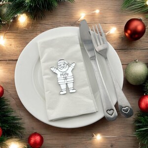 May include: A festive Christmas table setting with a white plate, napkin, and silverware. A silver Santa Claus place card holder with the name "Milly" is on the napkin. Red and green ornaments and pine branches decorate the wooden table.