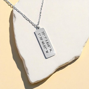Puede incluir: Un collar con colgante rectangular plateado y cadena fina. El colgante muestra las coordenadas "55° 7' 3.816" N, 1° 30' 40.03" W" en negro. La joya se exhibe sobre una superficie blanca texturizada.