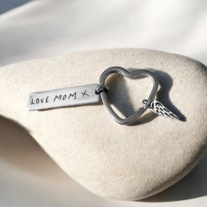 May include: A silver keychain with a heart-shaped design and a small wing charm. The keychain includes a rectangular tag with the inscription "LOVE MOM X". The heart is approximately 1.5 inches wide.