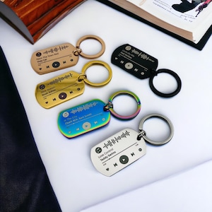 May include: Five different metal keychains with a music player design. Each keychain has a different song title and artist engraved on it. The keychains are in gold, black, rainbow, and silver.
