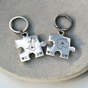 Puede incluir: Dos llaveros plateados con forma de piezas de rompecabezas. Cada pieza tiene un diseño de mapa con un corazón, las iniciales "E" y "B", y un símbolo de corazón. Los llaveros están unidos a anillos plateados.