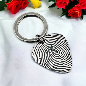 Puede incluir: Llavero de plata con un dije en forma de púa de guitarra con un diseño de huella digital en blanco y negro.