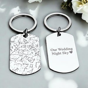 May include: A set of two silver keychains with a dog tag style design. One keychain features a constellation pattern and the other keychain has the text "Our Wedding Night Sky" with a heart symbol.