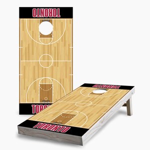 May include: A wooden basketball-themed cornhole game with a black and red "TORONTO" logo on the front. The game board features a wood grain design with a basketball court painted on it.