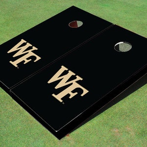 May include: Black cornhole game set with a gold "WF" logo on each board. The boards are set up on a green grassy surface.