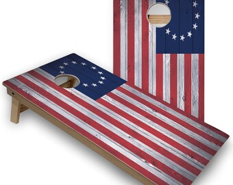 May include: A wooden cornhole game set with a red, white, and blue American flag design. The flag features 13 stars in a circle on a blue field. The game is made of wood and has a rustic look.