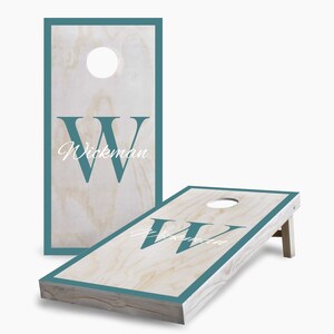 May include: A wooden cornhole set with a teal border and a white "W" and the name "Wickman" on each board. The boards are set up for play with one board standing upright.