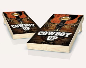 May include: Two wooden cornhole boards with a rustic brown background and a brown acoustic guitar with a black pickguard. The boards have the text "COWBOY UP" in white with a white star.