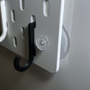 Op de afbeelding: Een wit prikbord met een zwarte plastic haak en een transparante zuignap. De haak is bevestigd aan het prikbord met een transparante plastic schroef.