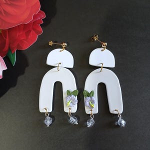 Puede incluir: Pendientes de cerámica blanca con forma de arco geométrico, acentuados con pequeñas flores moradas y cuentas de cristal transparentes.
