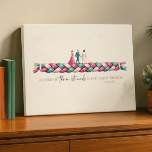 May include: A canvas print featuring a watercolor illustration of a braided rope in shades of pink and blue. Three figures walk across the rope. The text reads: "A cord of three strands is not easily broken."
