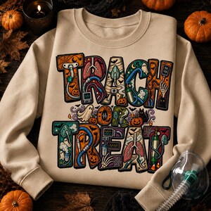 May include: A beige crewneck sweatshirt featuring the text "TRACH OR TREAT" in large, blocky letters. The letters are filled with colorful medical illustrations including lungs, oxygen masks, tubing, and Halloween motifs like pumpkins and ghosts.