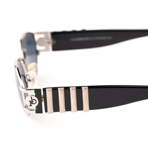 May include: Close-up of black sunglasses with silver accents. The sunglasses have a rectangular shape with a silver frame and black temples. The temples feature silver vertical stripes and a logo on the side.