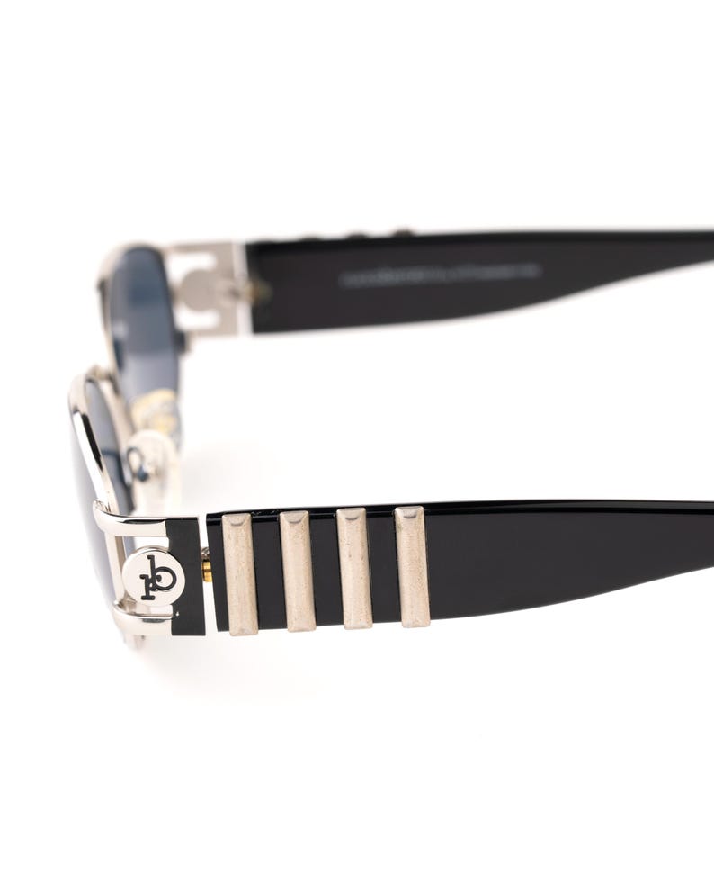 May include: Close-up of black sunglasses with silver accents. The sunglasses have a rectangular shape with a silver frame and black temples. The temples feature silver vertical stripes and a logo on the side.