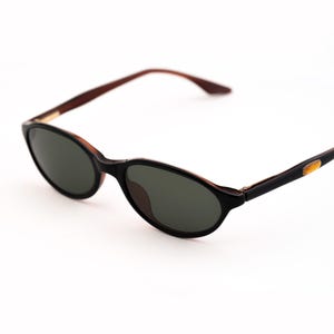 May include: Oval-shaped sunglasses with a black frame and dark lenses. The temples are brown and black, with a gold accent near the hinge. The sunglasses are set against a white background.