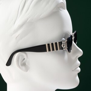 May include: Black sunglasses with silver and gold accents are displayed on a white mannequin head. The sunglasses have a rectangular frame with dark lenses. The temples feature vertical gold stripes and a silver emblem. The background is a dark green.