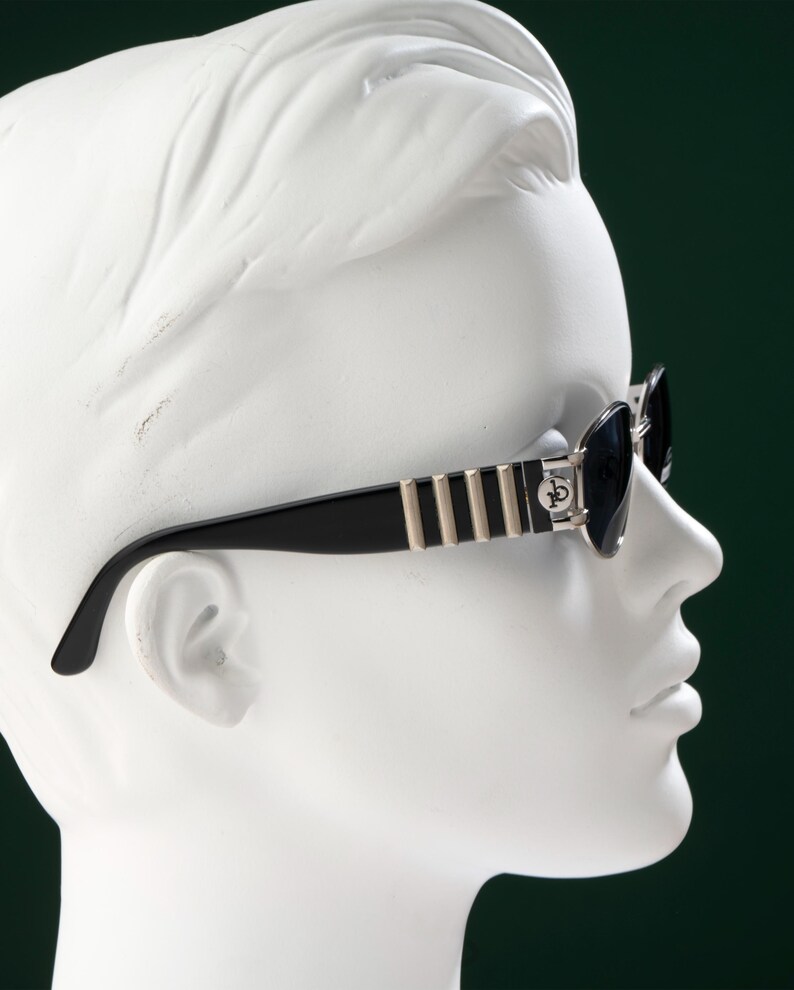 May include: Black sunglasses with silver and gold accents are displayed on a white mannequin head. The sunglasses have a rectangular frame with dark lenses. The temples feature vertical gold stripes and a silver emblem. The background is a dark green.