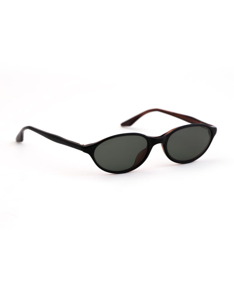 May include: Black oval sunglasses with dark lenses. The frames are black with brown arms. The sunglasses are set against a white background.