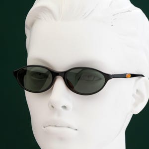 May include: Oval-shaped sunglasses with dark lenses and a black frame. The temples feature a small gold-colored accent. The sunglasses are displayed on a white mannequin head against a dark green background.
