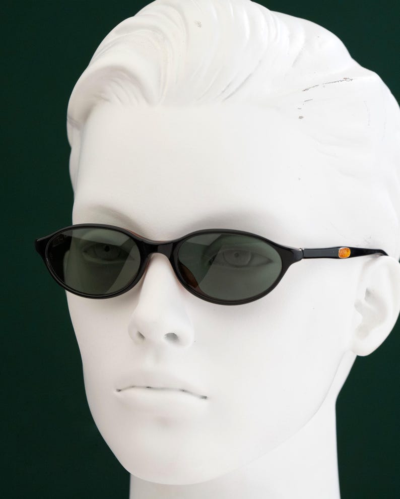 May include: Oval-shaped sunglasses with dark lenses and a black frame. The temples feature a small gold-colored accent. The sunglasses are displayed on a white mannequin head against a dark green background.