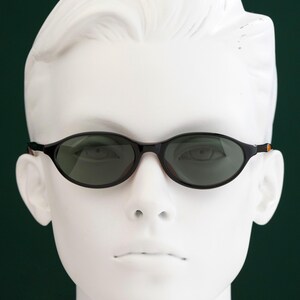 May include: Black oval sunglasses with dark lenses are displayed on a white mannequin head against a dark green background. The sunglasses have a thin frame with a touch of orange on the temples.