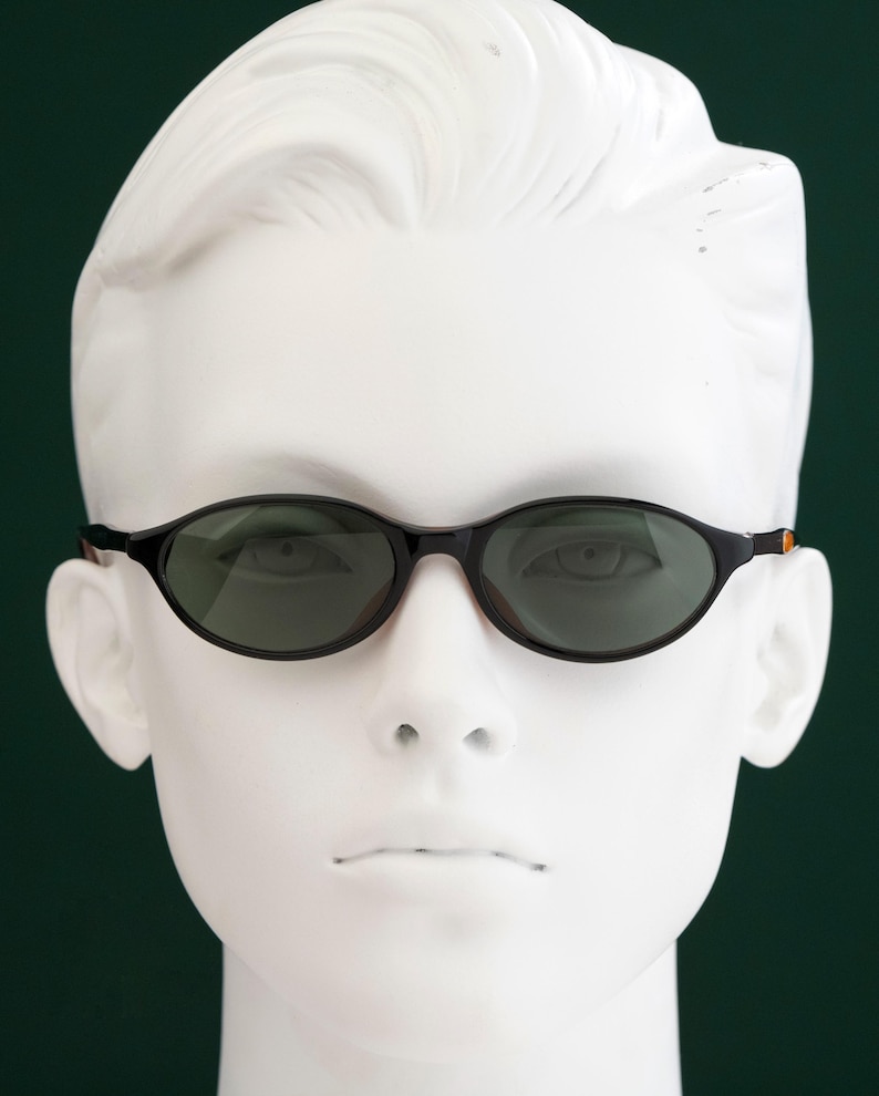 May include: Black oval sunglasses with dark lenses are displayed on a white mannequin head against a dark green background. The sunglasses have a thin frame with a touch of orange on the temples.