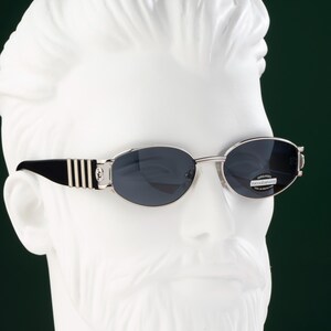 May include: Oval-shaped sunglasses with a silver frame and dark lenses. The temples are black with silver stripes and a decorative emblem. The sunglasses are displayed on a white bust against a dark green background.