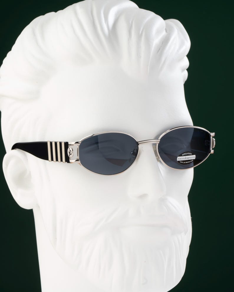 May include: Oval-shaped sunglasses with a silver frame and dark lenses. The temples are black with silver stripes and a decorative emblem. The sunglasses are displayed on a white bust against a dark green background.