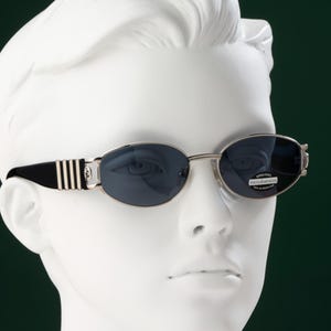 May include: Oval-shaped sunglasses with dark blue lenses and silver frames. The temples are black with silver stripes. The sunglasses are displayed on a white mannequin head against a dark green background.