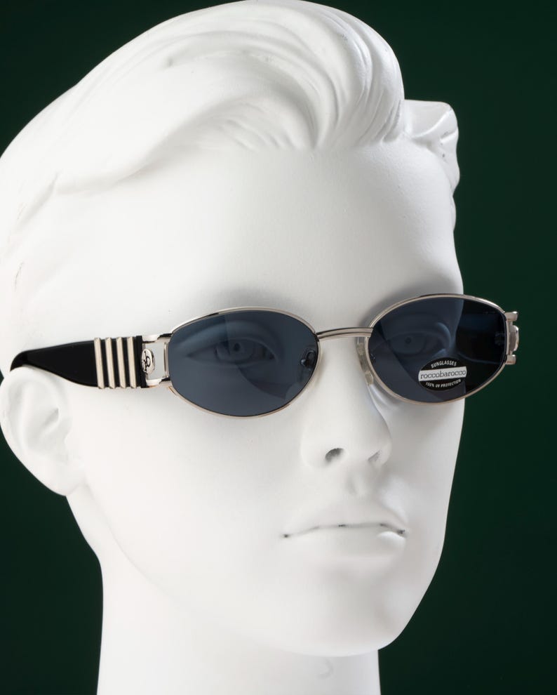 May include: Oval-shaped sunglasses with dark blue lenses and silver frames. The temples are black with silver stripes. The sunglasses are displayed on a white mannequin head against a dark green background.