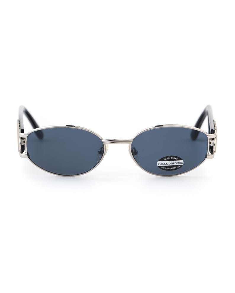 May include: Oval-shaped sunglasses with a silver frame and dark blue lenses. The sunglasses have a small sticker on the right lens with the text "ROCCOBAROCCO" and "100% UV PROTECTION". The temples are black.