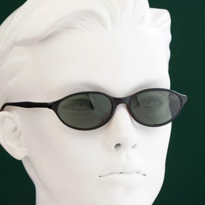 May include: Black oval sunglasses with dark lenses. The sunglasses have a thin, black frame and are displayed on a white mannequin head against a dark green background. The sunglasses are a classic style.