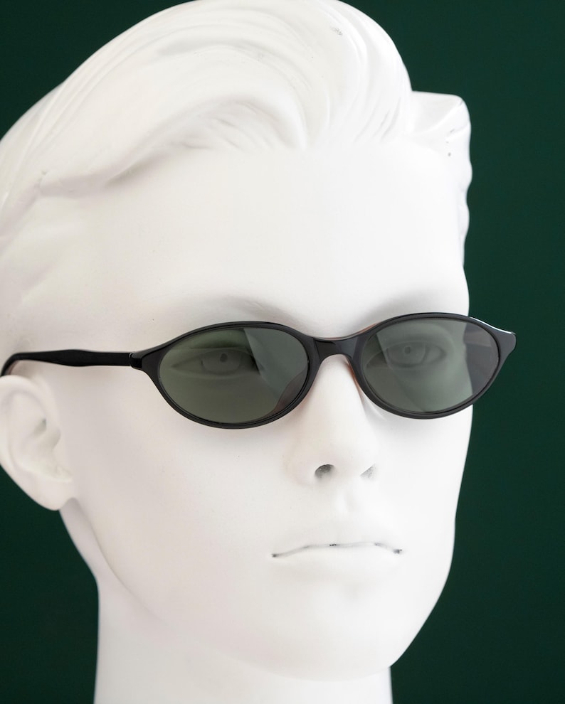 May include: Black oval sunglasses with dark lenses. The sunglasses have a thin, black frame and are displayed on a white mannequin head against a dark green background. The sunglasses are a classic style.