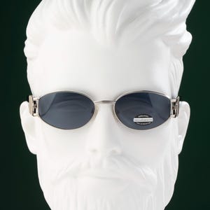 May include: Oval-shaped sunglasses with a silver frame and dark lenses. The sunglasses are displayed on a white mannequin head. The lenses have a small sticker with the text "roccobarocco".