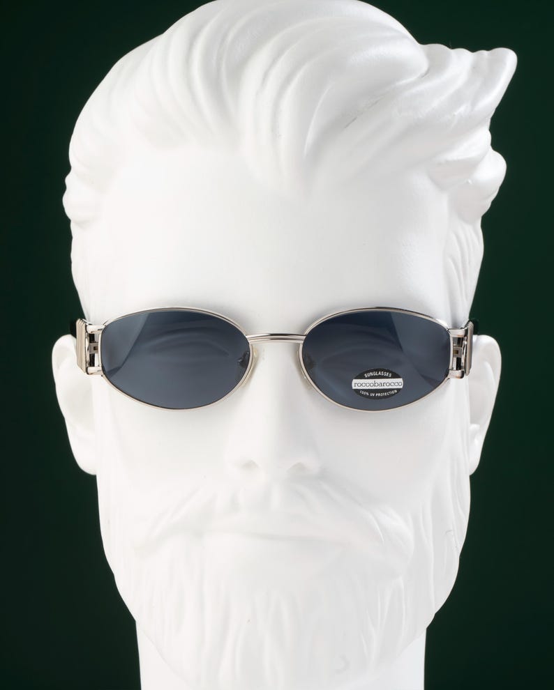May include: Oval-shaped sunglasses with a silver frame and dark lenses. The sunglasses are displayed on a white mannequin head. The lenses have a small sticker with the text "roccobarocco".