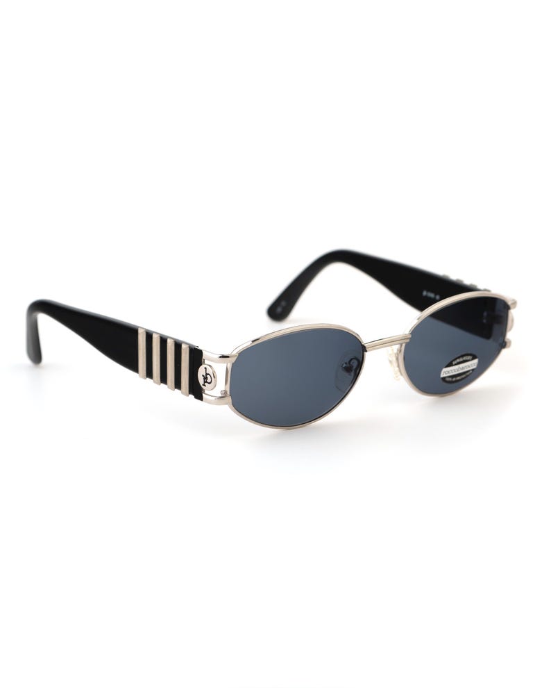 May include: Oval-shaped sunglasses with a silver frame and dark blue lenses. The temples are black with silver stripes and a logo. The sunglasses have a classic, retro design.